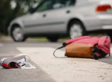 Close up photo of backpack and shoe on the ground next to a silver car, alluding to a person being struck by the car