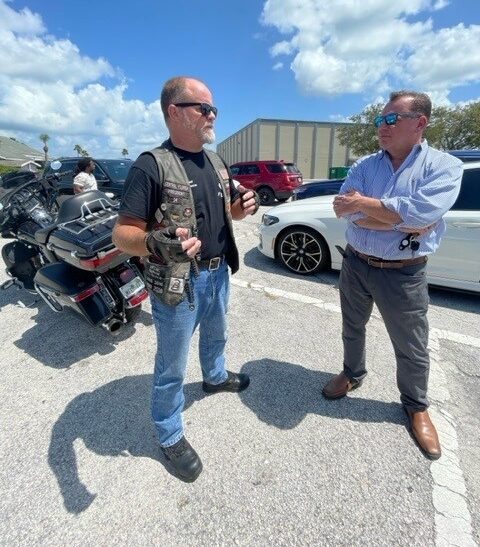 Attorney Rick Martindale in blue shirt with arms crossed talking with local nonprofit organization leader for MFC MC in front of harley davidson motorcycle